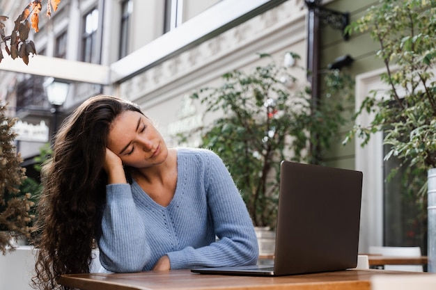 Уставшая молодая женщина делает перерыв на работе, спит и отдыхает за ноутбуком на рабочем месте из-за сверхурочной работы Спящая девушка с ноутбуком на столе на открытом воздухе в кафе