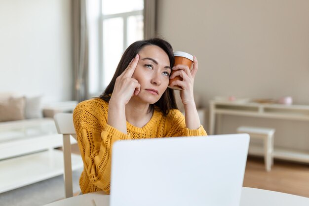 Tired young woman looking away and holding cup of coffee near head in office on blurred foreground