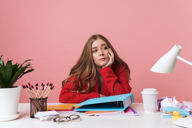 Photo a tired young woman sit at the table indoors isolated over pink wall with folder.