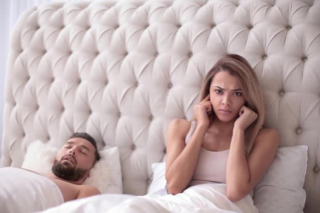 Tired young woman sitting in bed with snoring husband