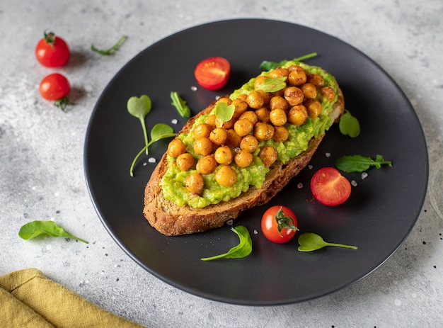 Toast met avocado, geroosterde kikkererwten, kerstomaatjes, kruiden op een donkere plaat gezond veganistisch voedsel grijs betonnen oppervlak