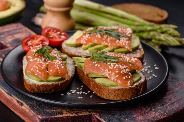 Toast sandwich with butter avocado and salmon decorated with arugula and sesame seeds on a black stone background