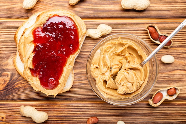 Toast sandwich with peanut butter. Spoon and jar of peanut butter, jam and peanuts for cooking breakfast on a brown wooden background. Creamy peanut paste flat lay.