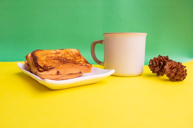 Toast On White Plate And Black Coffee Mug With Spruce Flower Decoration On Yellow And Green Paper Background. Toast And Black Coffee For Breakfast. Horizontal Photo