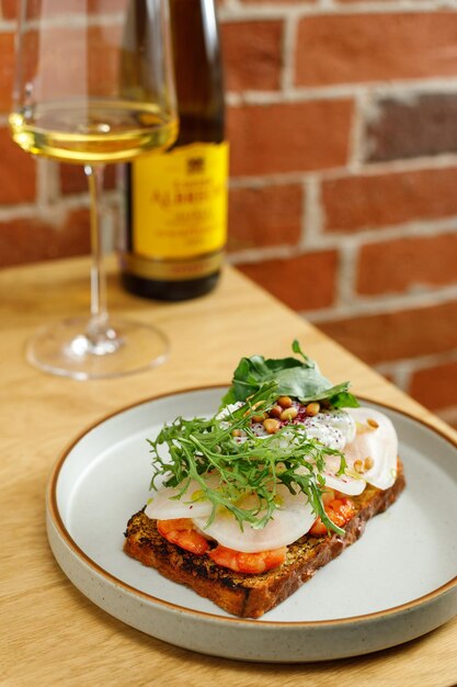 Toast with grilled shrimps and vegetables poached egg and herbs on top served in restaurant Wine bottle and glass on background