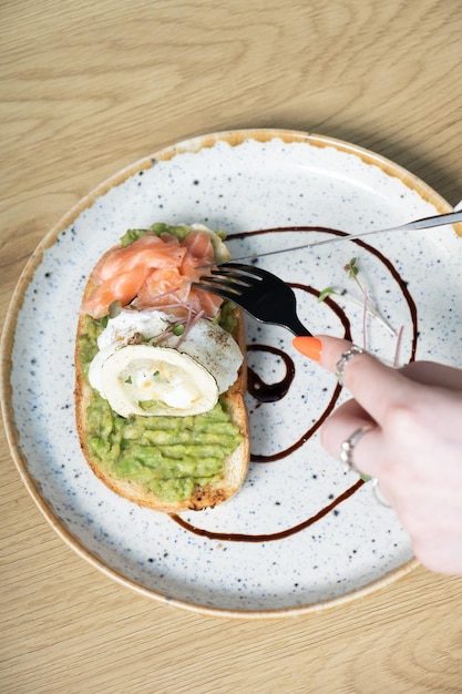 Toast with salmon and a poached egg on an avocado spread top view Female hands with a fork