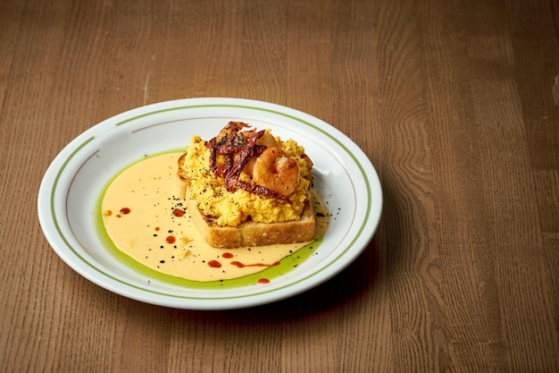 Toast with scrambled eggs shrimp and hollandaise sauce in a plate on a wooden background