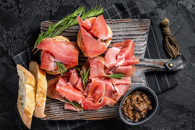 Toast with tomatoes and cured Slices of jamon serrano ham prosciutto crudo parma on wooden board with rosemary Black background Top view