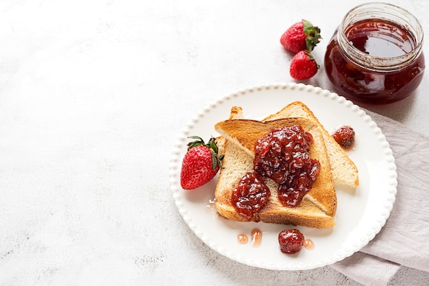 Toasted bread slices with strawberry jam and a jar of jam on white wooden background