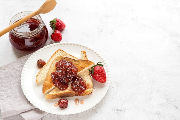 Toasted bread slices with strawberry jam and a jar of jam on white wooden background