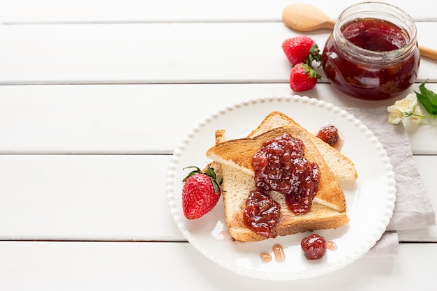 Toasted bread slices with strawberry jam and a jar of jam on white wooden background