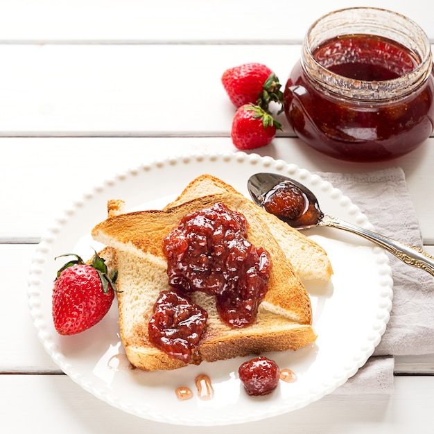 Toasted bread slices with strawberry jam and a jar of jam on white wooden background