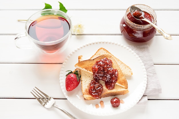 Toasted bread slices with strawberry jam and a jar of jam on white wooden background