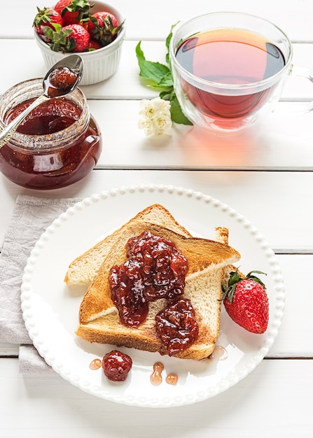 Toasted bread slices with strawberry jam and a jar of jam on white wooden background