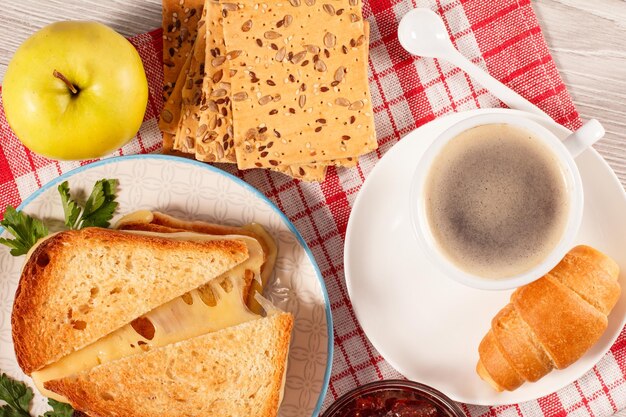 Toasted slices of bread with cheese, apple, cookies with whole grains of sunflower seeds, cup of coffee and croissant on saucer and glass bowl with strawberry jam on red napkin. Top view.