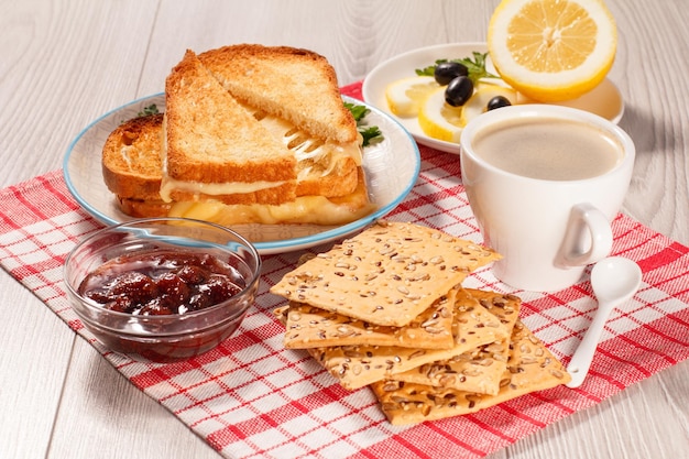 Toasted slices of bread with cheese, cookies with whole grains of sunflower seeds, cut lemon on plate, cup of coffee and glass bowl with strawberry jam on red napkin.