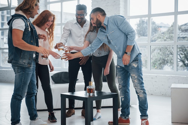 Toasting for the bright future. Young people having good time in the office. Smiling and drinking with windows at background