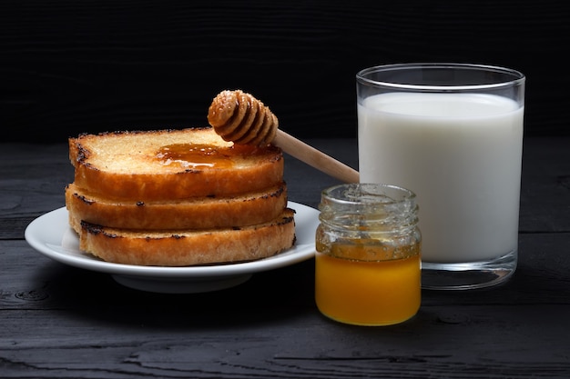 Toasts on a white plate, a glass of milk and a jar of honey with a honey spoon