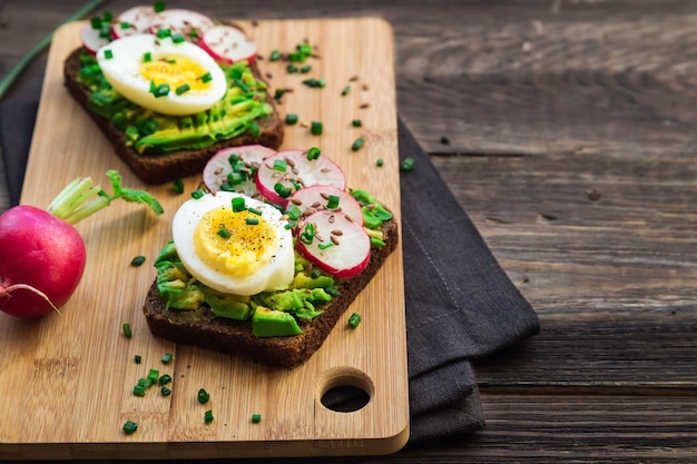 Toast con avocado, uova, ravanello, cipolla verde e semi di lino su fondo di legno rustico.