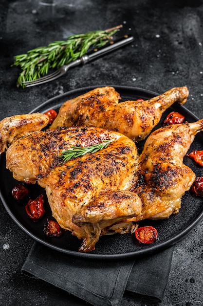 Tobacco whole chicken on plate with herbs and tomato. Black background. Top view.