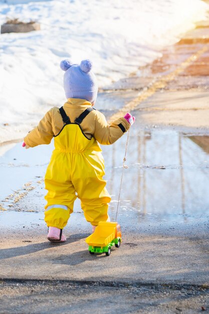 Toddler 23 years old dressed in yellow waterproof clothes runs through puddles with a toy truck tied on a rope