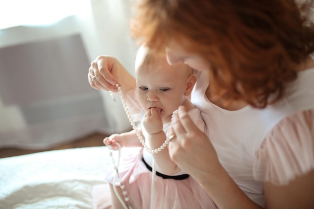 Photo toddler baby in a dress with beads with a red-haired mother playing on a bed in a real interior