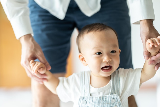 사진 걷는 법을 배우고 가정에서 아버지와 어머니와 함께 어린 아이와 함께 가족의 지원을 받는 유아 아기는 도움으로 걷고 아빠의 손을 잡고 행복한 어린 시절 도움 보살핌을 받는 첫 걸음을 내딛습니다.