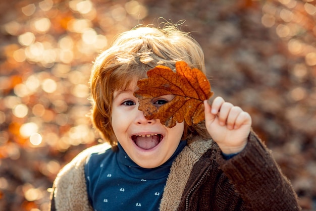 Foto ragazzo del bambino nel parco d'autunno. il bambino si copre gli occhi con una foglia d'acero gialla nel parco autunnale. bambini che camminano nel parco d'autunno. ragazzo carino che gioca con le foglie di acero all'aperto. ritratto di bambino autunnale.