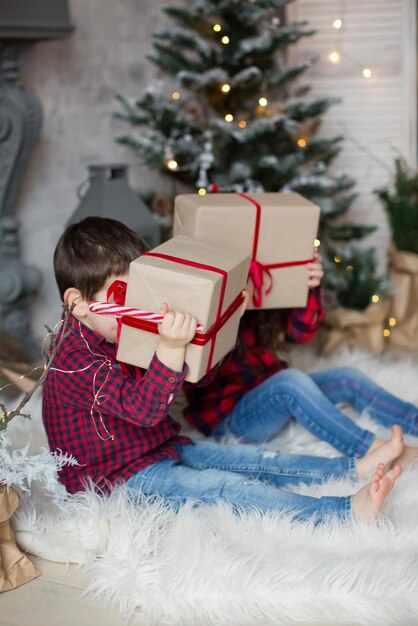 빨간 셔츠를 입은 유아 소년과 소녀는 밝은 크리스마스 인테리어로 크리스마스 트리 근처에 선물 상자를 들고 있습니다. 귀여운 크리스마스 아이들