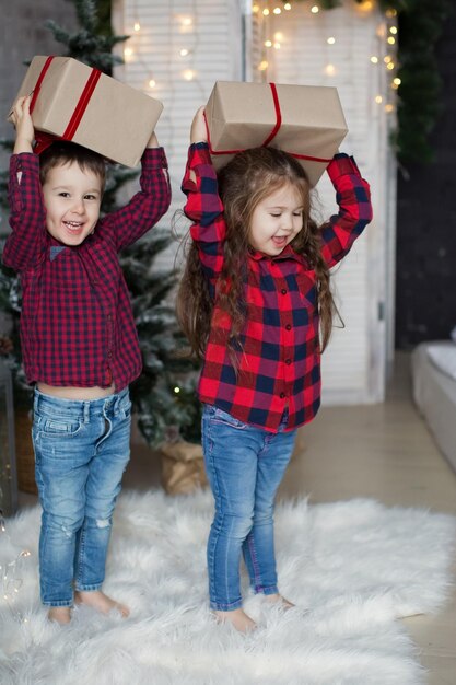 Toddler boy and girl in red shirts hold gift boxes near Christmas tree in light Christmas interior Cute Christmas kids