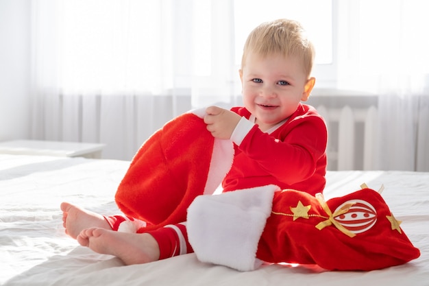 Малыш мальчик в красном костюме с рождественским носком, сидя на белой кровати у себя дома. солнечное утро.