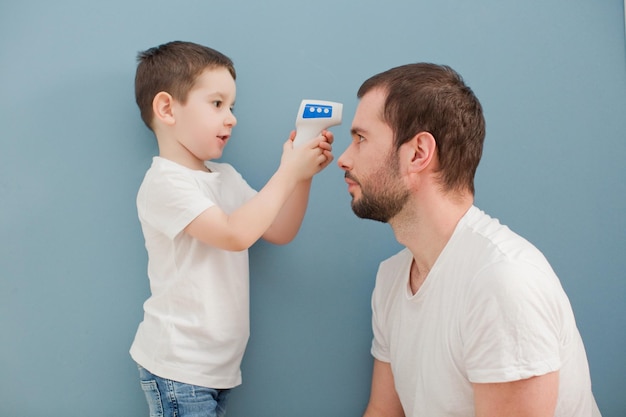 Toddler boy is measuring his fathers body temperature using infrared thermometer on a blue background