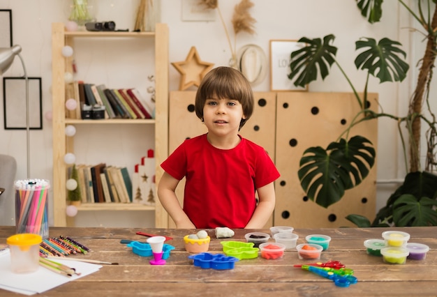 Il bambino in una maglietta rossa si siede a un tavolo con forniture creative e guarda la parte anteriore della stanza