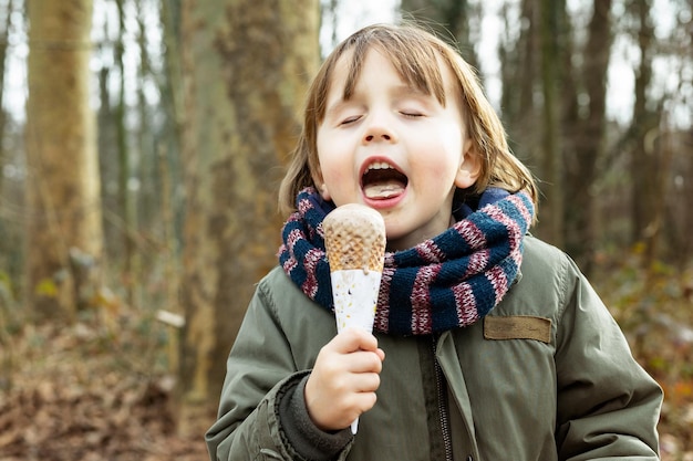 Toddler boy wearing warm jacket and scarf eating chocolate ice cream in waffle cone with pleasue outdoor in cold weather