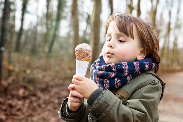 Toddler boy wearing warm jacket and scarf eating chocolate ice cream in waffle cone with pleasue outdoor in cold weather