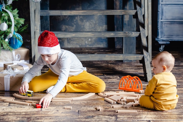 Мальчики в шляпе Санты строят железную дорогу и играют с игрушечным поездом под елкой. Украшенный дом для зимнего отдыха. Дети с рождественскими подарками. Рождественское время.