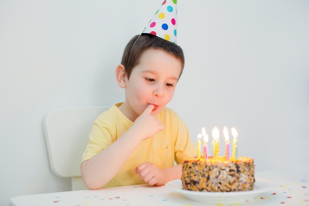 흰색 배경에 촛불을 불고 생일 케이크 근처에 앉아 다채로운 생일 모자에 유아 백인 소년