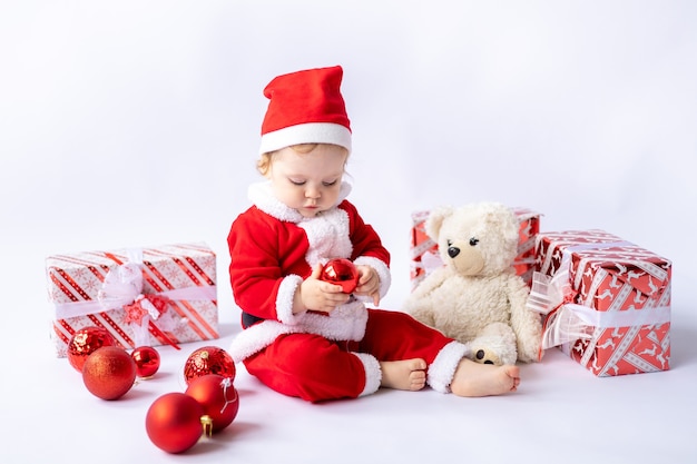 白い背景の上に座ってクリスマスプレゼントを保持しているサンタの衣装を着た幼児の子供新年