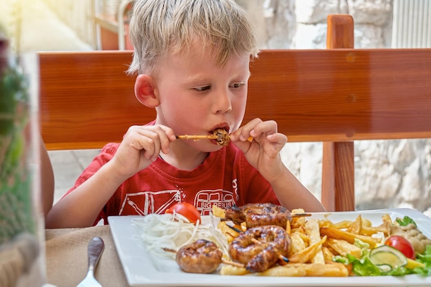 幼児は新鮮な空気のレストラン テラスでフライド ポテトと野菜と一緒に肉を食べる