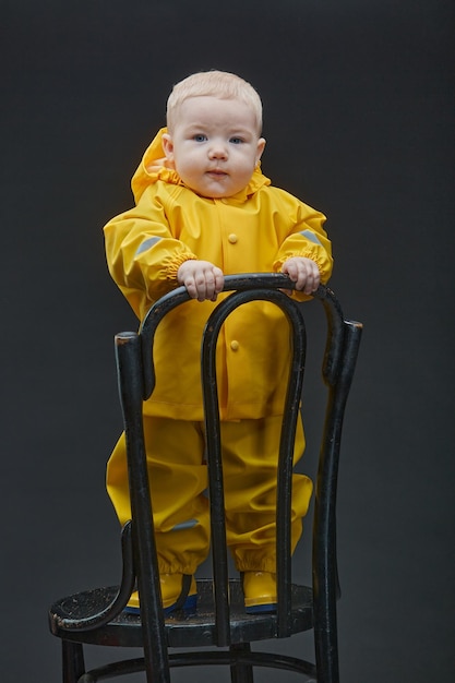 A toddler in fireman's suit stands on chair and looks into camera