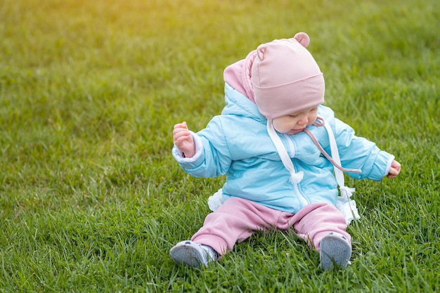 太陽に照らされた緑の草の上に座る暖かい服を着た幼児の女の子