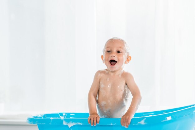 Toddler kid taking bath and smiling in blue plastic baby bathtub