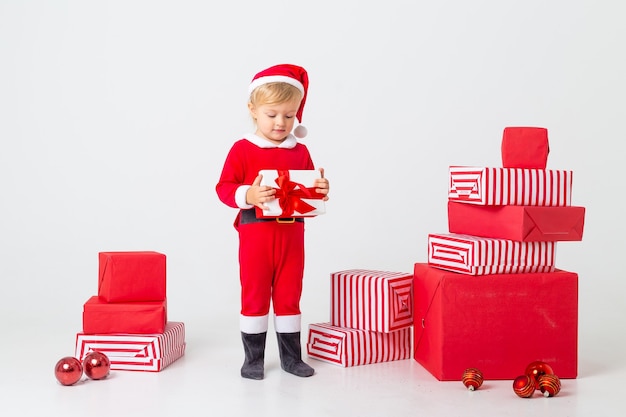 Un bambino in costume da babbo natale si trova su uno sfondo bianco accanto a scatole regalo per natale. concetto di natale, spazio di testo