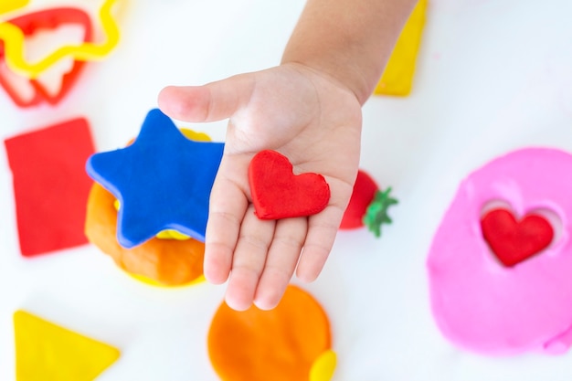 유아는 흰색 테이블에 색된 Plasticine에서 조각합니다. 어린 아이의 손이 유색 플라스틱 조각을 짜냅니다. 어린이의 창의력, 교육용 게임, 훌륭한 운동 능력. 플라 스티 신 심장