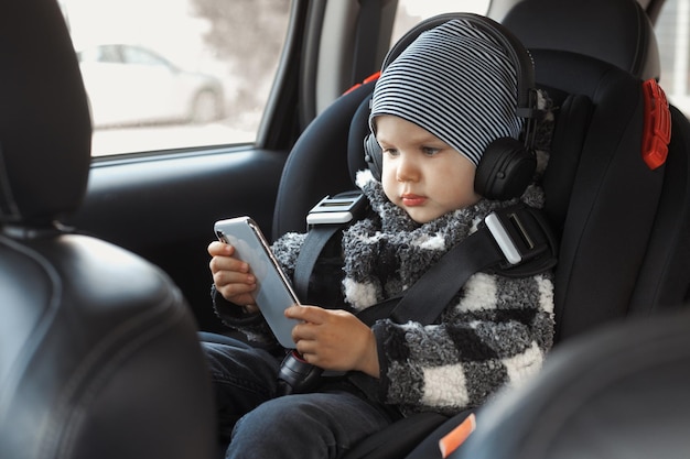 Toddler sits in a child car seat and watches cartoons with\
headphones and holds a mobile phone