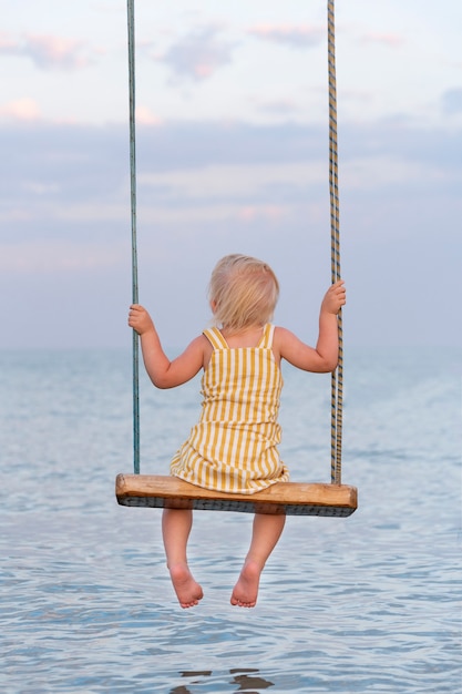 Toddler sitting on rope swing over the water. Back view. Vertical frame. Happy childhood.