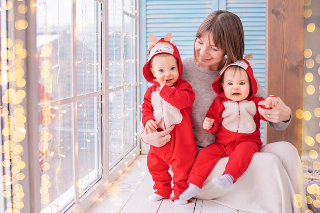 赤いトナカイのサンタクロースの衣装を着た幼児の双子は、花輪の窓の背景に母親と一緒に家に座っています