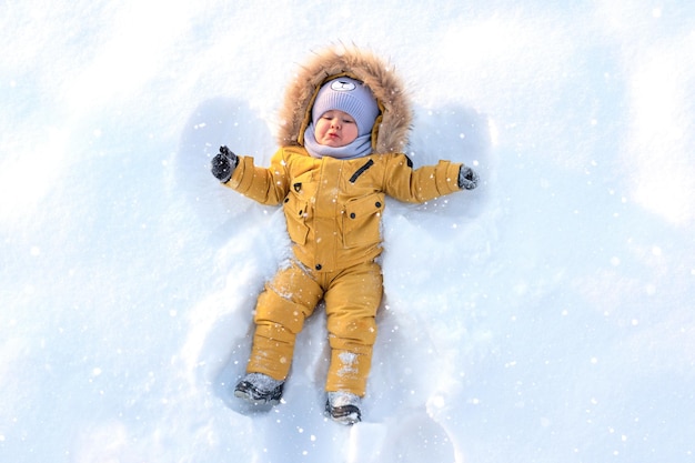 Toddler in yellow winter clothes lies on the snow and makes a snow angel.