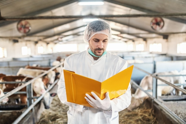 Toegewijde ongeschoren dierenarts in beschermend uniform met haarnetje dat in de schuur staat en de testresultaten bekijkt.