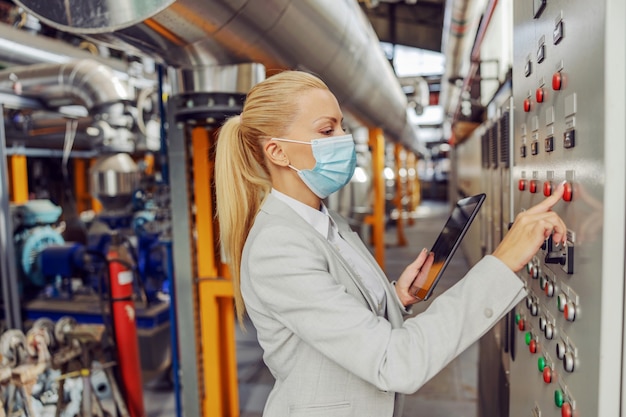 Toegewijde vrouwelijke blonde supervisor met gezichtsmasker die in verwarmingsinstallatie naast dashboard staat, instellingen aanpast en tablet vasthoudt tijdens coronavirus-pandemie.